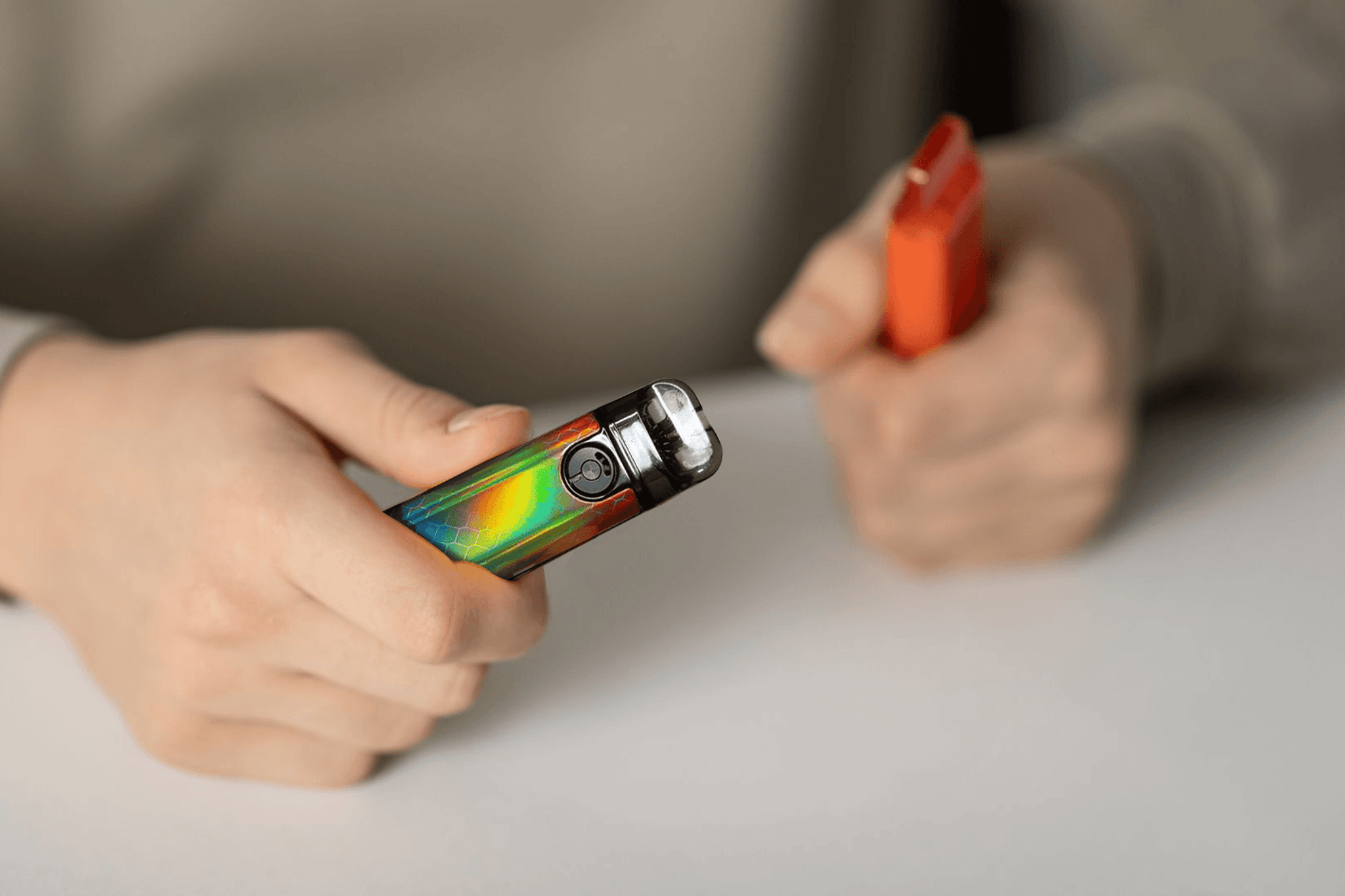 Close-up of hands holding a rainbow refillable vape and an orange disposable vape.